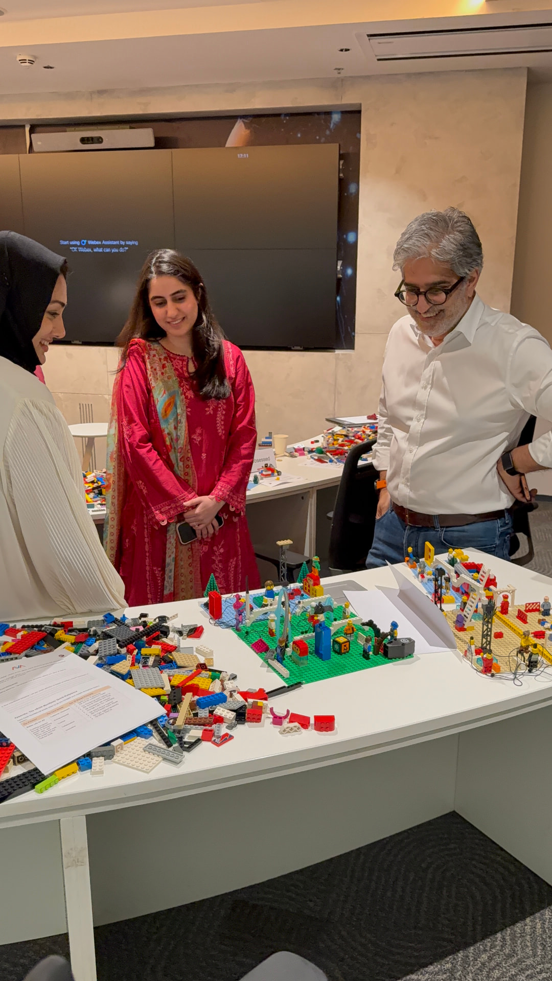 Three participants — a woman in a magenta ethnic dress, a woman in a white hijab, and an older man in glasses — collaborating around a white table covered in loose LEGO bricks during an AVA LEGO AdVentures workshop